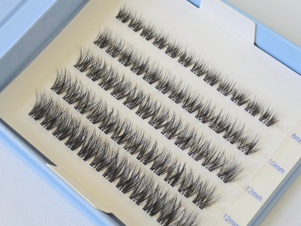 A close-up of false eyelash clusters arranged in rows on a white card inside a light blue box. The card shows measurements in millimeters along the right edge.