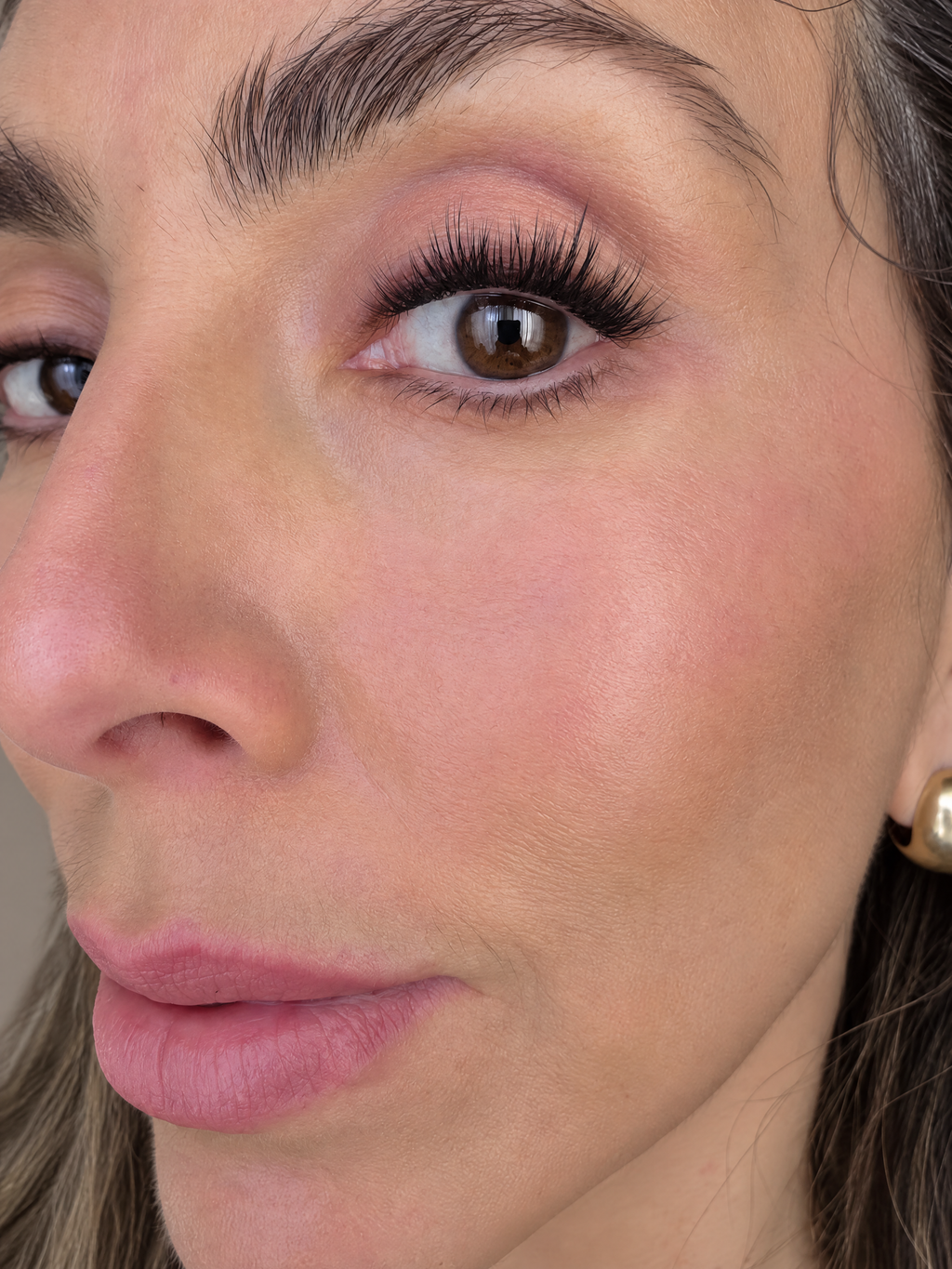 Close-up of a woman's face showing eyelash application, featuring long black false eyelashes, pink eyeshadow, and a small gold stud earring.