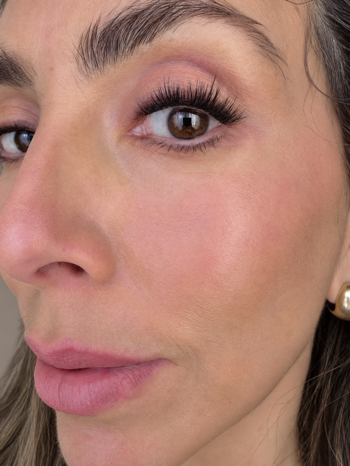 Close-up of a woman's face showing eyelash application, featuring long black false eyelashes, pink eyeshadow, and a small gold stud earring.