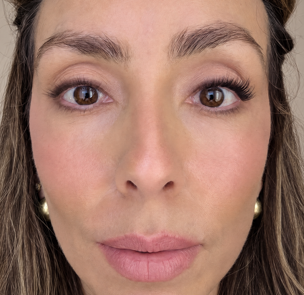 Close-up of a woman's face wearing a false lashes in one eye, showing her eyes, nose, and lips against a neutral beige background.
