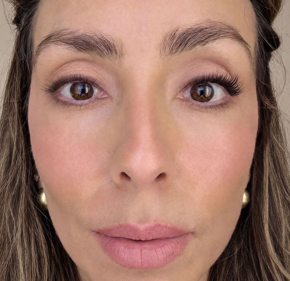 Close-up of a woman's face wearing a false lashes in one eye, showing her eyes, nose, and lips against a neutral beige background.